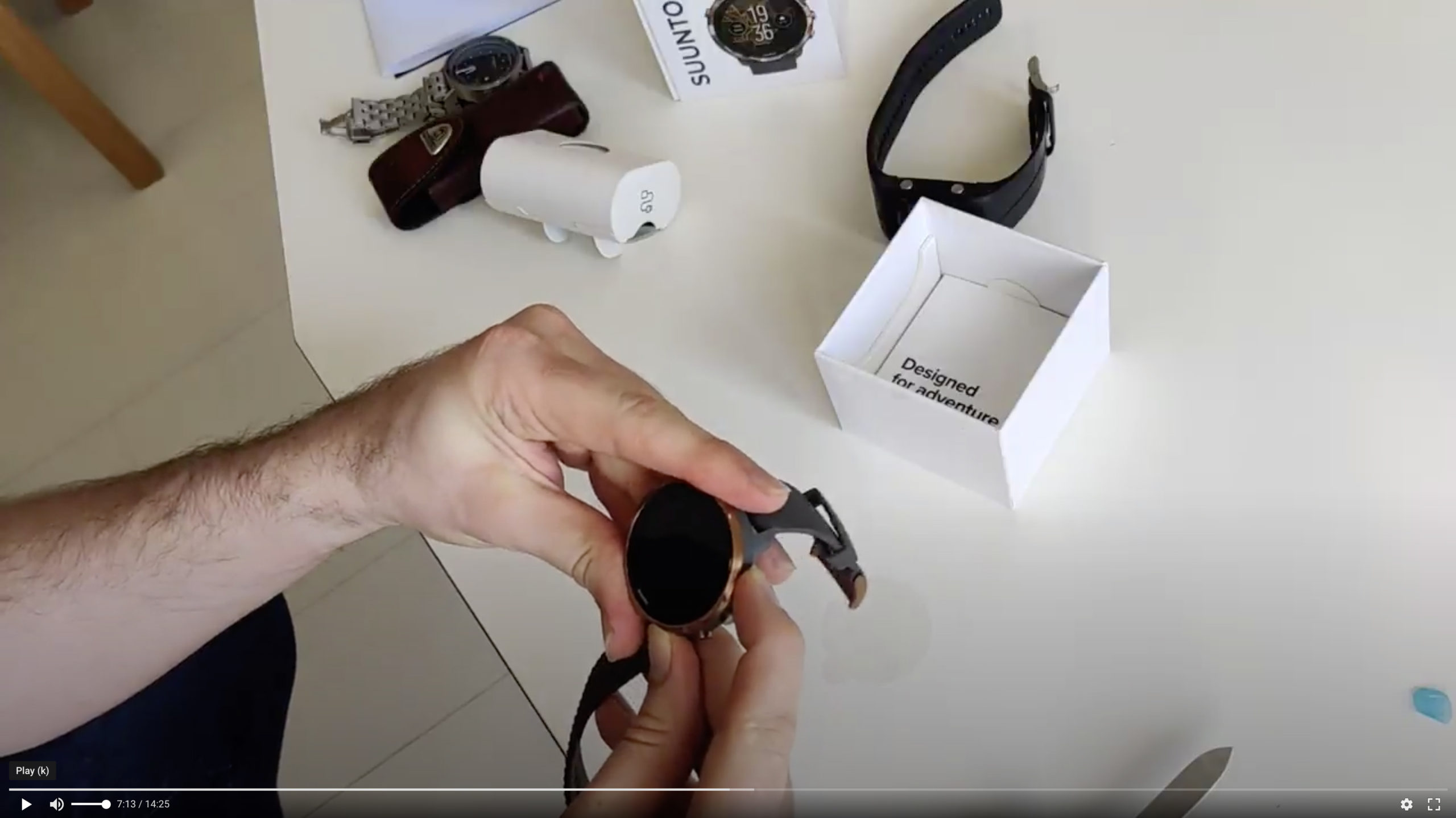 Screen capture of a video that shows the Suunto 7 unboxing. The photo shows two hands holding a watch. On the background you can see a desk and a watch package that has been opened.
